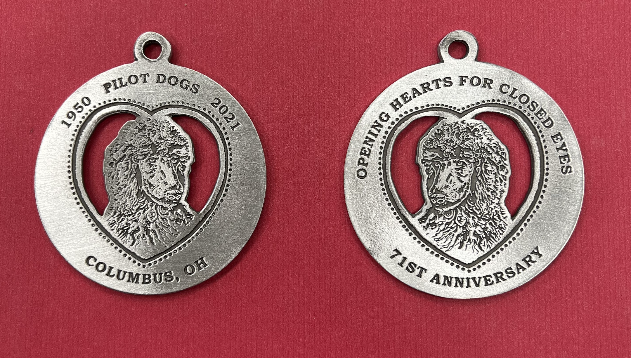 Two Ornaments to show both sides of the Standard Poodle Pewter Ornament. One side has 1950 Pilot Dogs 2021 above the engraved poodle headshot and Columbus, OH below. The other side has Opening hearts for closed eyes above the engraved poodle headshot and 71st Anniversary below.