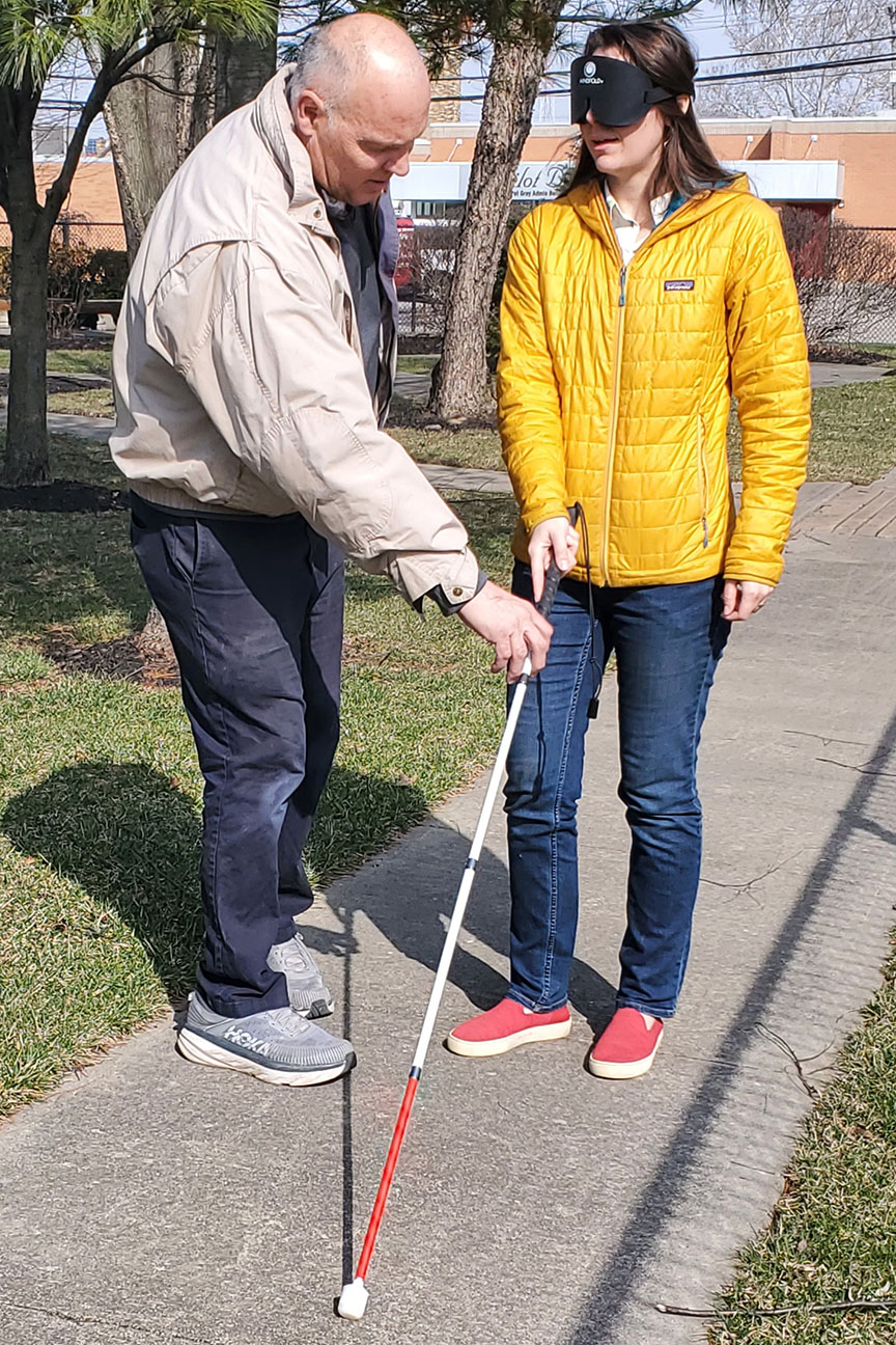 A student in a yellow jacket stands blindfolded on the sidewalk while the instructor adjusts how she holds her white cane.