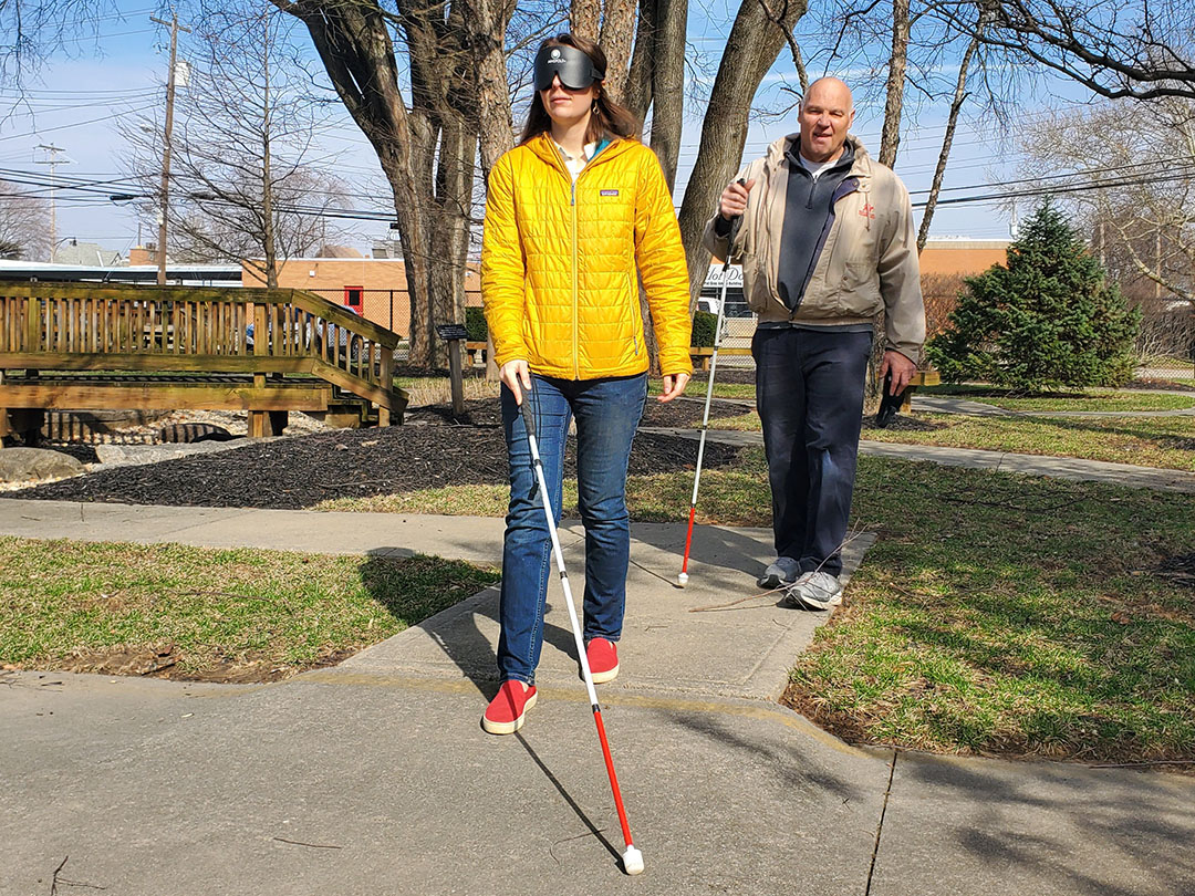 A student with a white cane is wearing a blindfold, walking on the sidewalk. Behind her is the instructor, also with a white cane.