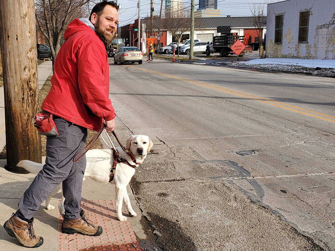 A trainer standing at the curb with a yellow Lab in harness on his left, looking to their right before crossing the street.