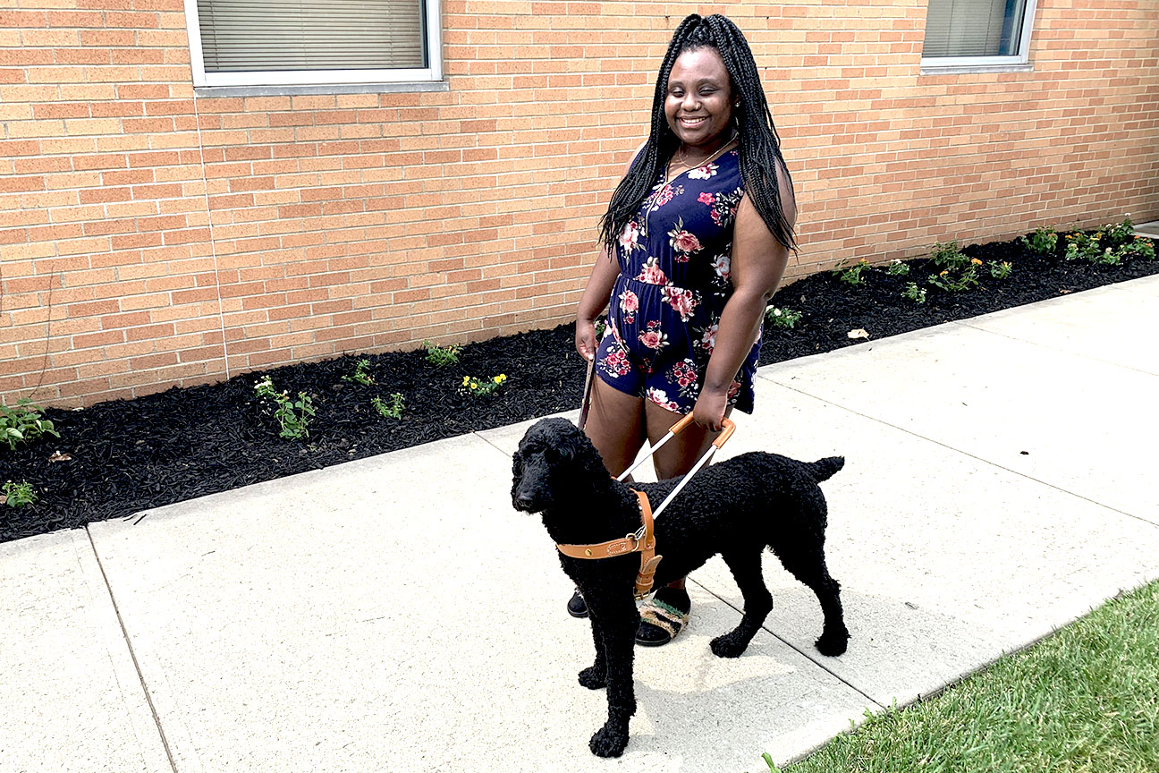 A student in a floral summer dress stands smiling on the sidewalk with a black standard poodle in harness on her left.